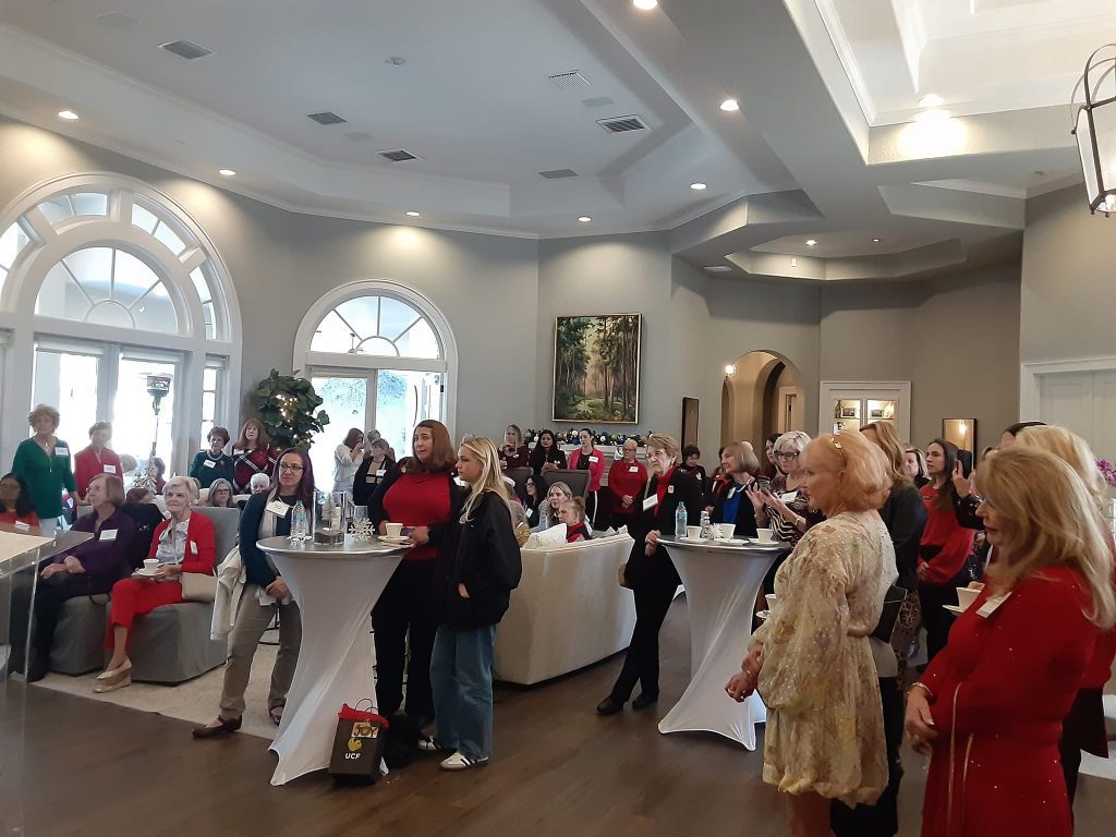 A group of people stands and sits in a well-lit room with arched windows, engaging in a social or networking event.