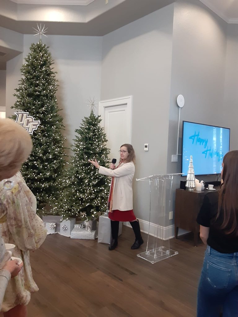 A woman with a microphone gestures near decorated Christmas trees, with people watching. A screen displays "Happy Holidays.