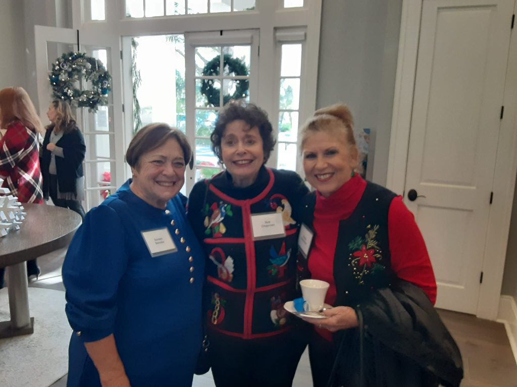 Three women in festive sweaters pose together at an indoor event, with one holding a cup. Christmas decorations are visible in the background.