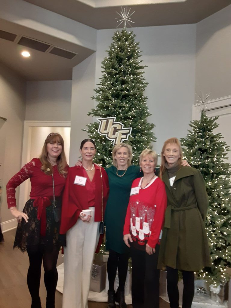 Five women standing in front of Christmas trees, dressed in holiday attire, with a "UCF" sign visible above them.