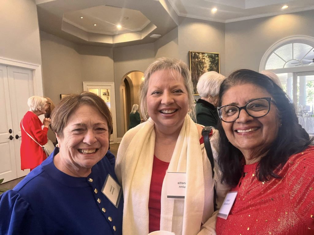 Three women smiling at a social event indoors. They are wearing name tags and standing close together, with other attendees visible in the background.
