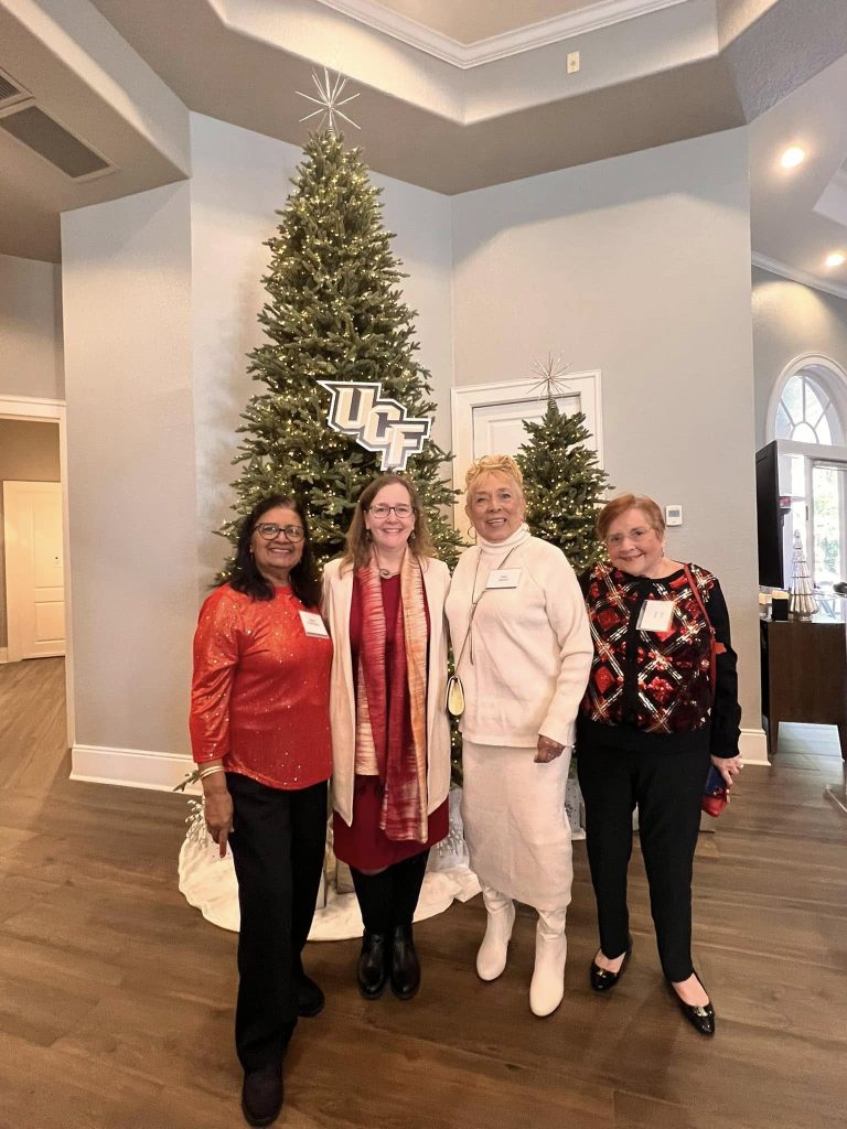 Four women stand smiling in front of decorated Christmas trees with a UCF sign, in a warmly lit room with a high ceiling.