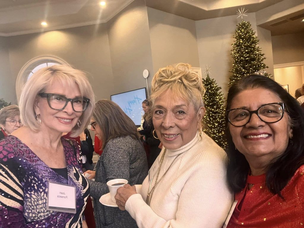 Three women smiling in a festive setting with Christmas trees in the background. One woman holds a cup of coffee. They are wearing name tags.