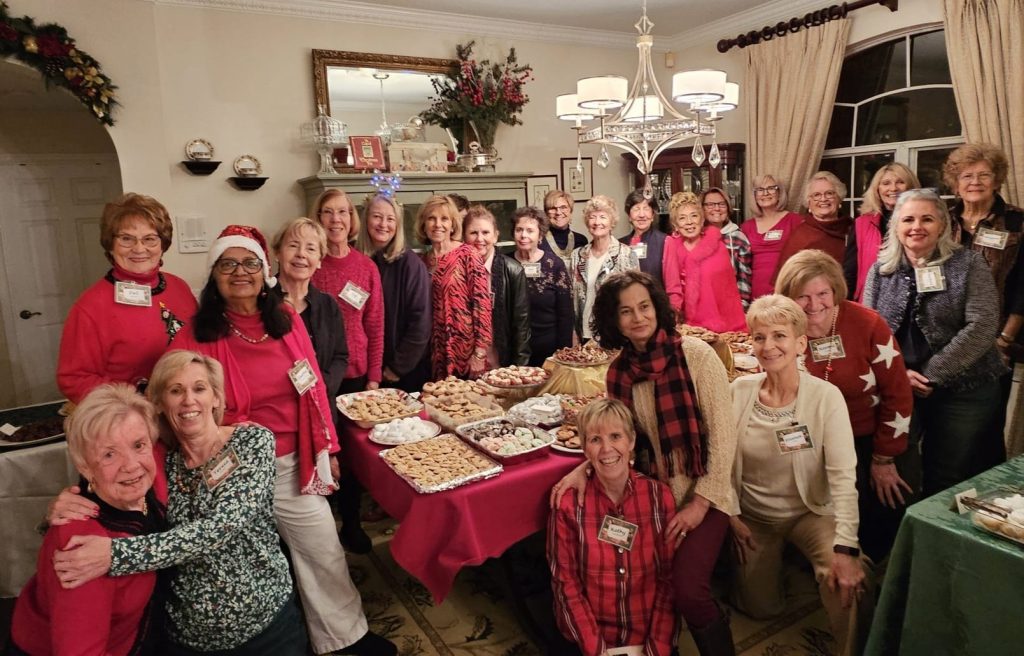 A group of women gather around a table filled with various desserts in a warmly decorated room.