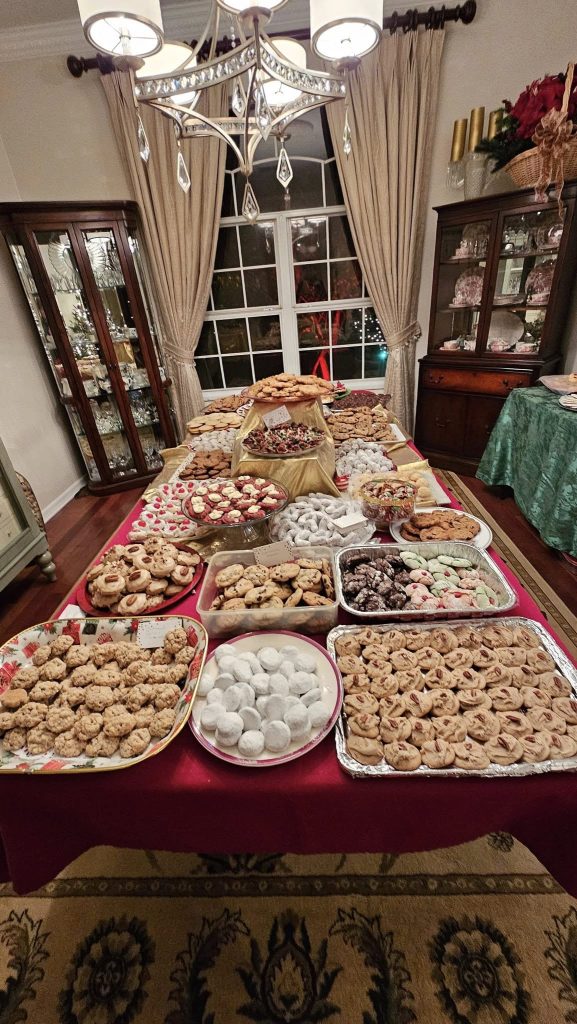 A table covered with a variety of cookies and pastries is set in a dining room with a chandelier overhead and curtains by the window.