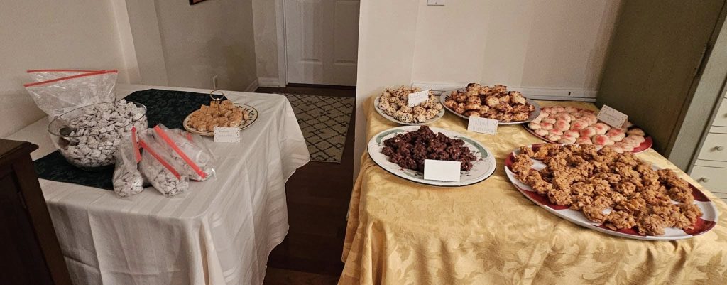 A table with various desserts including cookies, pastries, and snacks on plates and in bags, set on tablecloths in a room with a closed door in the background.