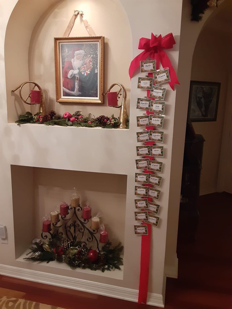 A festive wall display with a framed Santa picture, holiday foliage, candles, and a red ribbon holding labeled cards.