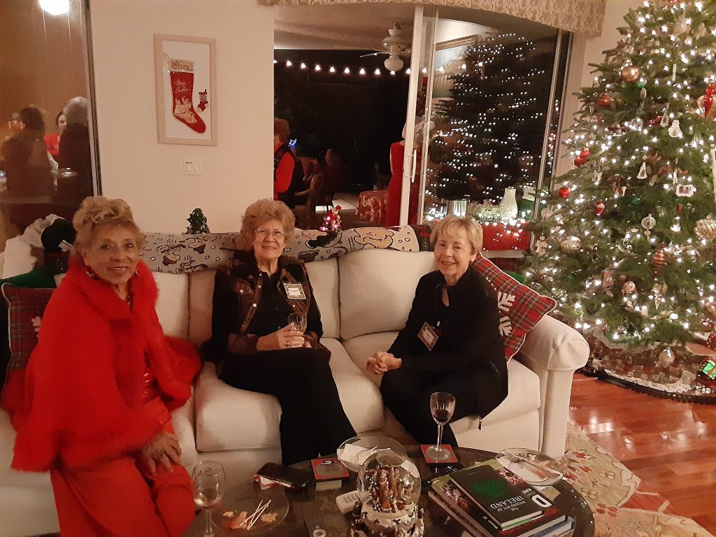 Three women sitting on a couch with wine glasses, surrounded by holiday decorations and a Christmas tree, in a warmly lit living room.