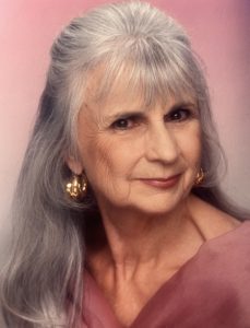 Elderly woman with long gray hair and gold earrings, wearing a pink top, poses against a pink background.