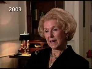 An older woman with styled hair speaks in a room with a lamp and bookshelf visible in the background. "2003" is written in the upper left corner.