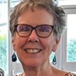 Woman with short hair and glasses, smiling, wearing earrings and a patterned top, standing indoors with windows in the background.