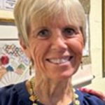 An older woman with short, light hair smiles while wearing a blue top and a gold necklace, standing in an indoor setting with framed pictures on the wall behind her.