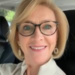 A woman with short blond hair and glasses smiles while sitting in a car. She is wearing a light-colored top and earrings.
