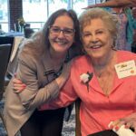 Two women smiling at an indoor event; one wears a blazer with a name badge, and the other wears a pink blouse with a corsage and a name tag reading "Ann O’Leary Smith.