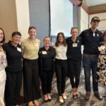 Seven women and one man stand in a line indoors, smiling at the camera. They are dressed in business casual and athletic attire, with name tags visible.
