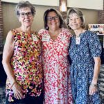 Three women stand together indoors, smiling at the camera. Each is wearing a patterned dress and a name tag. There are brick walls and a “Hall of Fame” sign in the background.