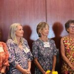 Six women stand side by side in front of wooden doors, wearing patterned dresses and name tags, looking in different directions.