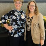 Two women stand side by side, smiling at the camera. One wears a floral top and name badge; the other wears glasses, a patterned blouse, and a beige blazer.