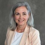 A woman with shoulder-length gray hair, wearing a light-colored blazer over a white top, smiles in front of a plain gray background.
