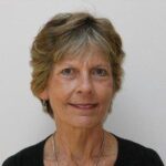 A woman with short, light brown hair and blue eyes is wearing a black top and a necklace, posing in front of a plain light background.
