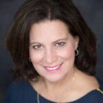 A woman with shoulder-length brown hair, wearing a dark top, gold hoop earrings, and a necklace, smiles at the camera against a dark background.