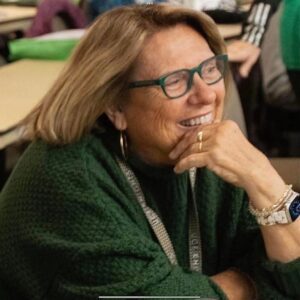 A woman with glasses and a green sweater sits at a desk, smiling with her hand resting on her chin.