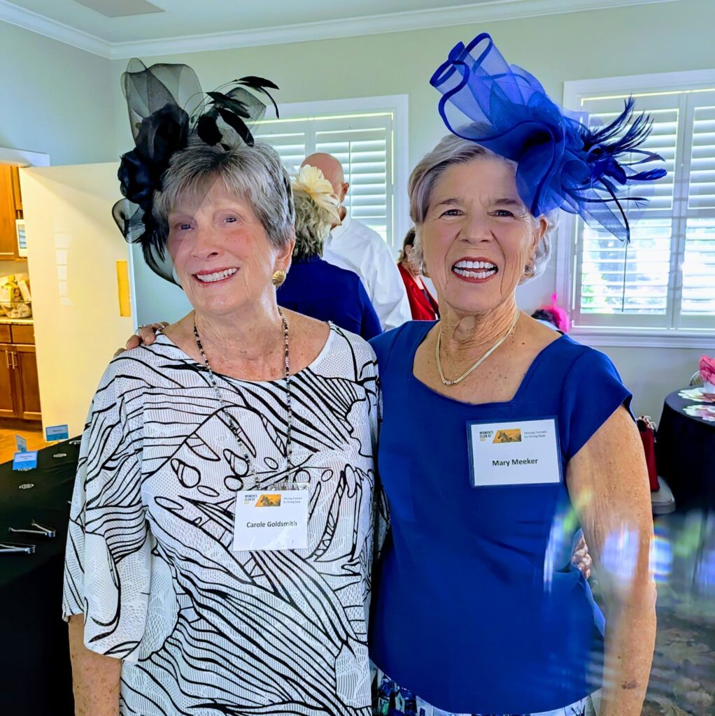 Two older women standing together indoors, both wearing decorative hats and name tags, smiling at the camera.