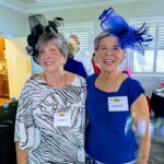 Two older women standing together indoors, both wearing decorative hats and name tags, smiling at the camera.