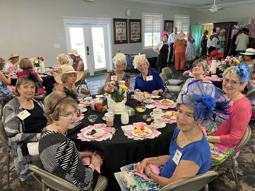 A group of women wearing hats sit around decorated tables with tea and snacks, participating in a social gathering in a bright indoor venue.