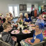 A group of women wearing hats sit around decorated tables with tea and snacks, participating in a social gathering in a bright indoor venue.