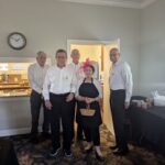 Five adults stand in a room near tables with food and drinks. One woman in the center wears a pink headpiece, while the four men wear white shirts. A clock is on the wall above them.