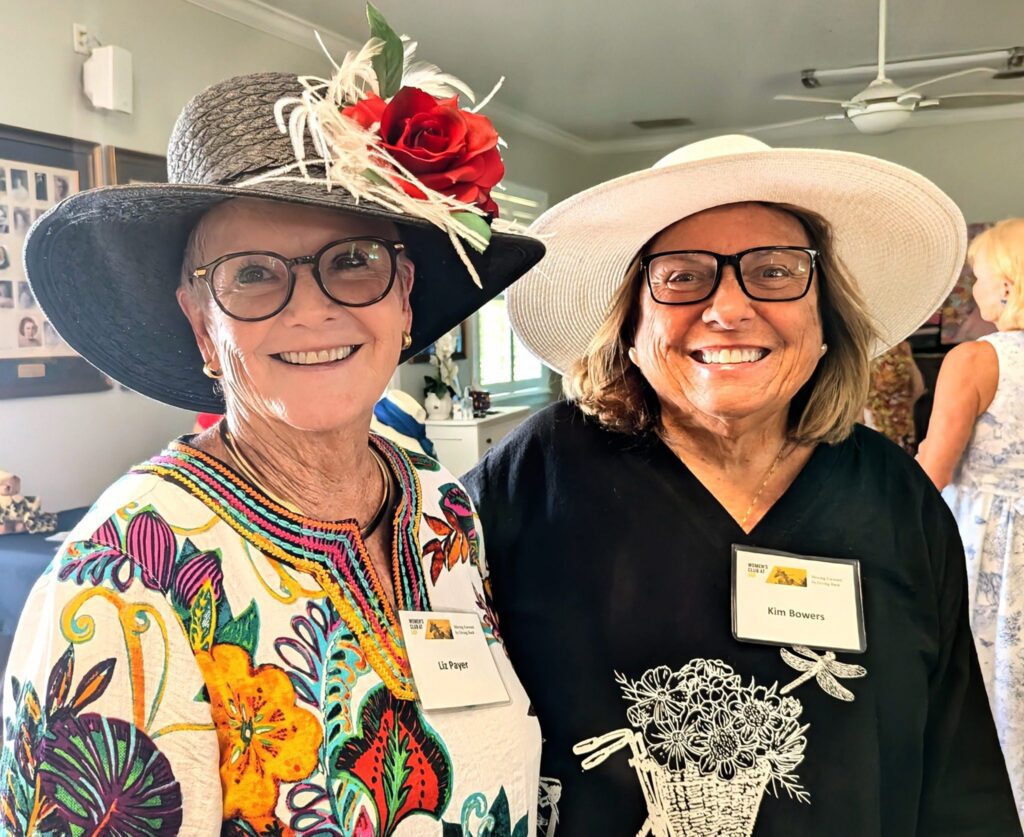 Two smiling women wearing large hats and name tags stand indoors at an event. One woman wears glasses and a colorful floral top, the other wears glasses and a black shirt with a floral design.