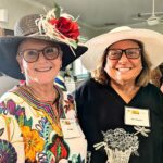 Two smiling women wearing large hats and name tags stand indoors at an event. One woman wears glasses and a colorful floral top, the other wears glasses and a black shirt with a floral design.