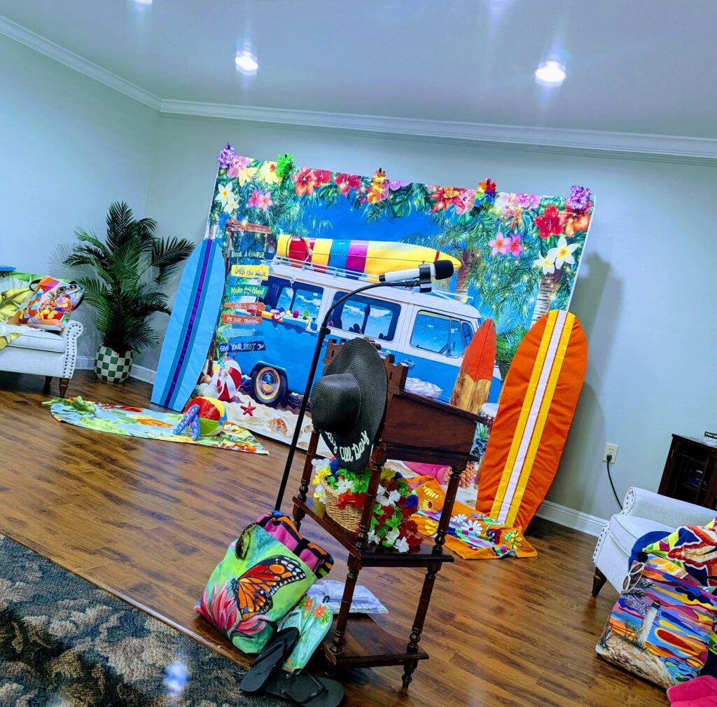 A brightly decorated room features a beach-themed backdrop with a van, surfboards, tropical props, and colorful accessories scattered on the floor and furniture.
