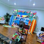 A brightly decorated room features a beach-themed backdrop with a van, surfboards, tropical props, and colorful accessories scattered on the floor and furniture.