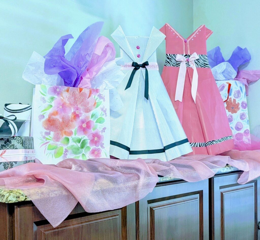 Gift bags decorated with tissue paper and dress-shaped designs sit on a counter with a light pink sheer fabric draped in front.