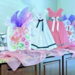 Gift bags decorated with tissue paper and dress-shaped designs sit on a counter with a light pink sheer fabric draped in front.