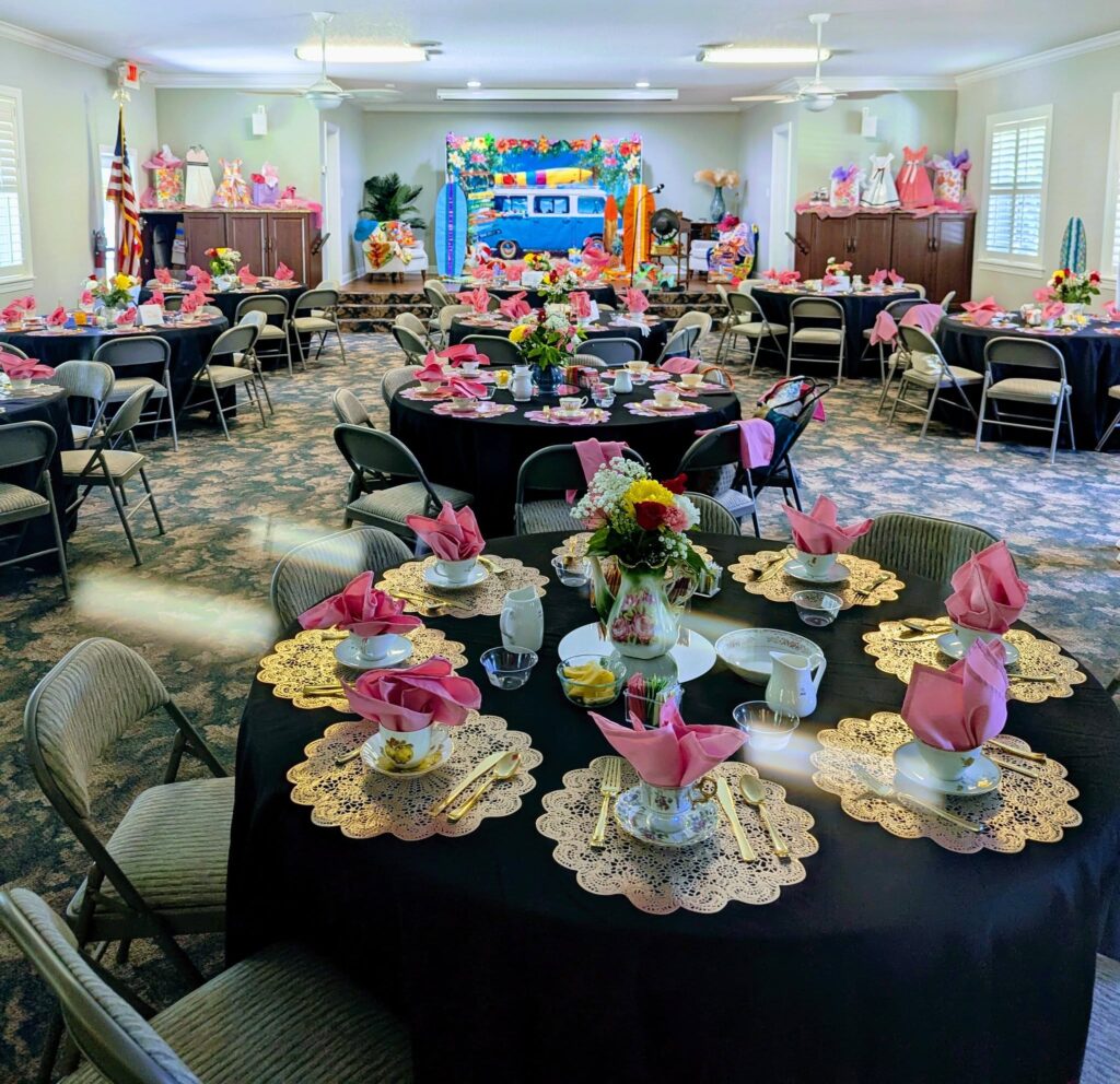 A banquet hall is set up with round tables covered in black cloth, pink napkins, floral centerpieces, and teacups, prepared for a formal tea party event.