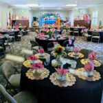 A banquet hall is set up with round tables covered in black cloth, pink napkins, floral centerpieces, and teacups, prepared for a formal tea party event.