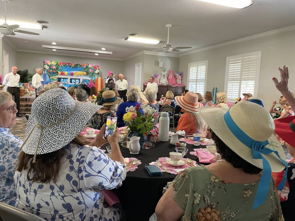 A group of women in hats sit at tables with tea settings, watching a small group of people standing and performing at the front of a decorated event room.