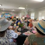 A group of women in hats sit at tables with tea settings, watching a small group of people standing and performing at the front of a decorated event room.
