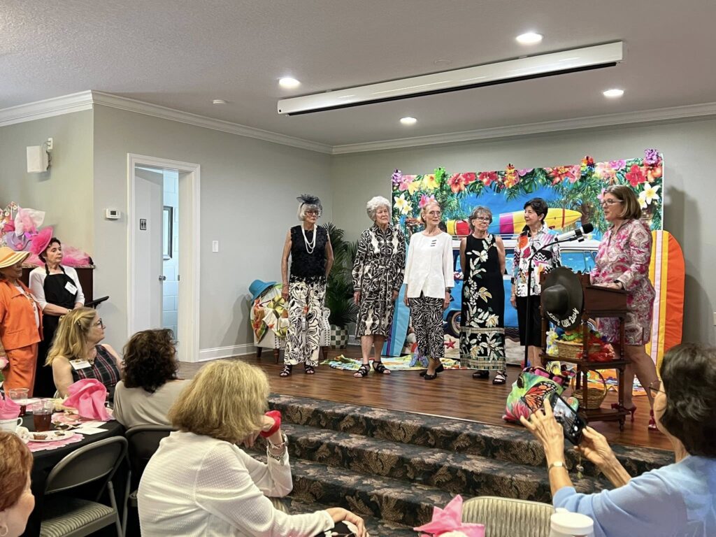 A group of women stand on a decorated stage in a room, with others seated and watching. The setting appears festive, with a beach-themed backdrop and colorful decorations.