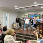 A group of women stand on a decorated stage in a room, with others seated and watching. The setting appears festive, with a beach-themed backdrop and colorful decorations.