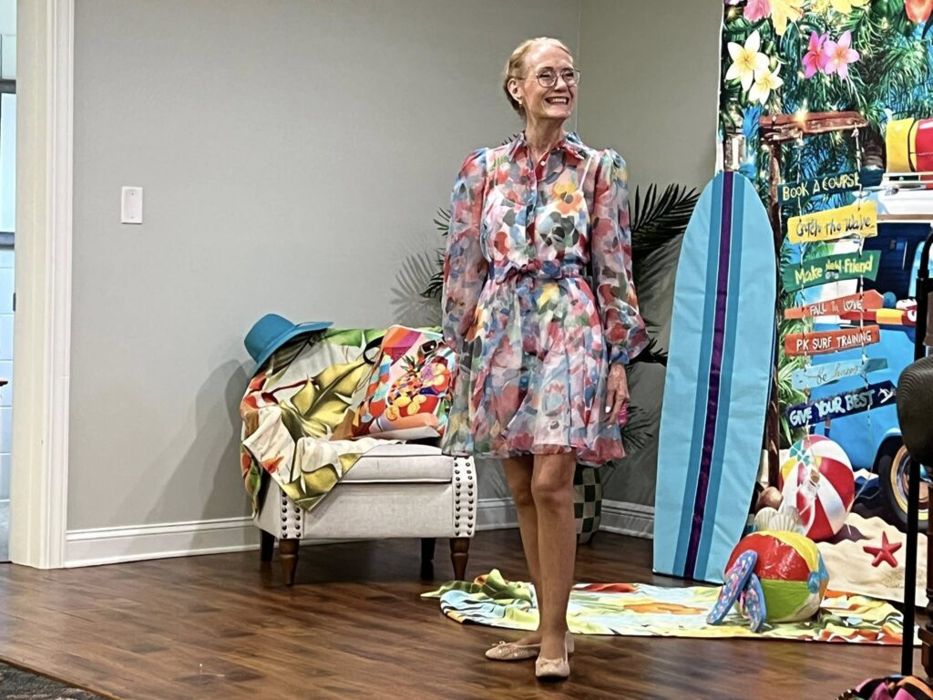 An older woman stands indoors wearing a colorful, floral dress and glasses, smiling. Behind her are beach-themed decorations, a chair, and a wooden floor.