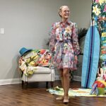 An older woman stands indoors wearing a colorful, floral dress and glasses, smiling. Behind her are beach-themed decorations, a chair, and a wooden floor.
