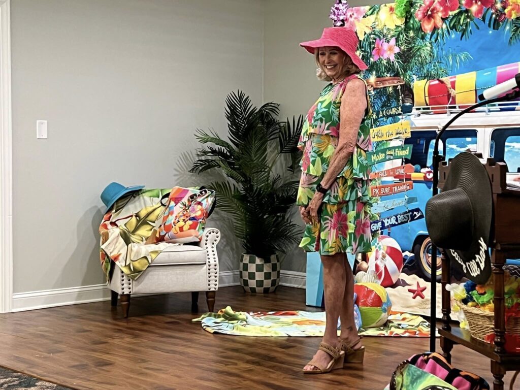 A woman in a floral dress and pink sunhat stands indoors near a tropical-themed backdrop and summer decorations.