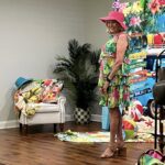 A woman in a floral dress and pink sunhat stands indoors near a tropical-themed backdrop and summer decorations.