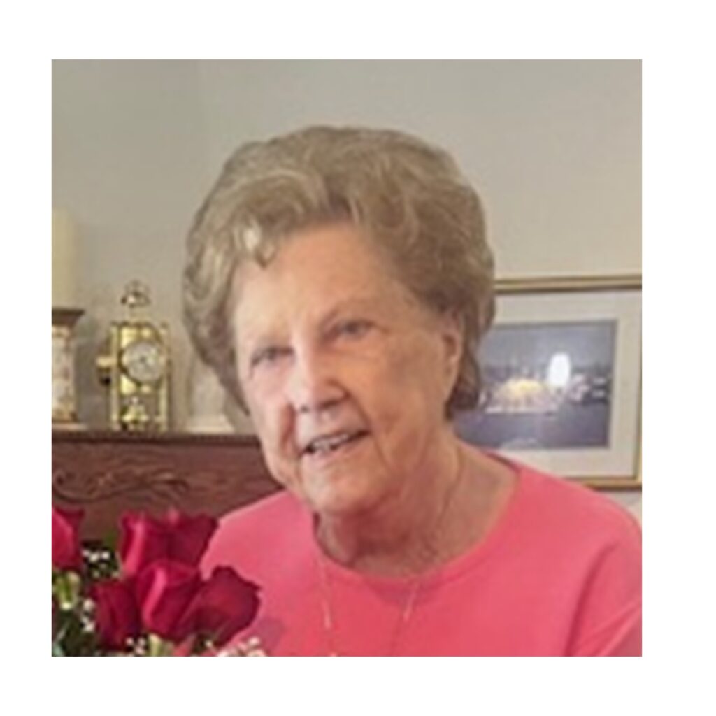 An elderly woman with short, curly hair wearing a pink top sits indoors, holding a bouquet of red roses. A framed picture and a clock are visible in the background.