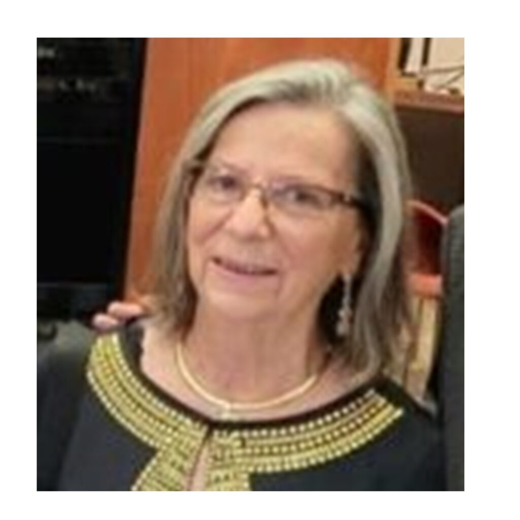An older woman with glasses and shoulder-length gray hair, wearing a black top with gold trim and a pearl necklace, smiles at the camera.