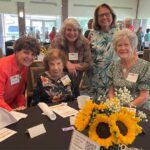 Five women pose and smile around a table with sunflowers at a social event held in a bright, spacious room filled with other attendees.